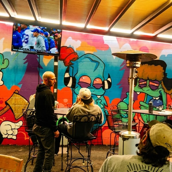 Patrons relax on an outdoor patio with colorful graffiti art, a TV, and patio heaters.