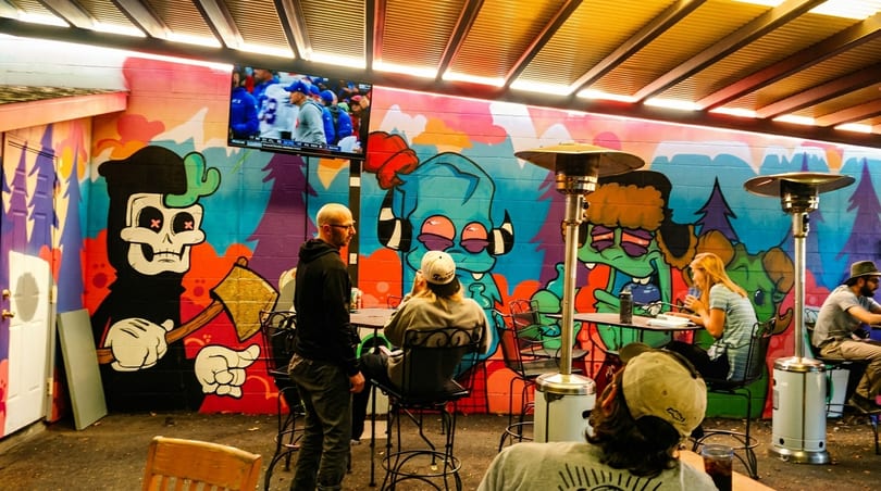 Patrons relax on an outdoor patio with colorful graffiti art, a TV, and patio heaters.