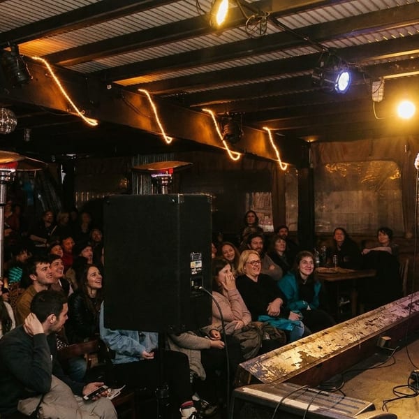 A comedian performs on stage for a laughing audience in a dimly lit venue with string lights.