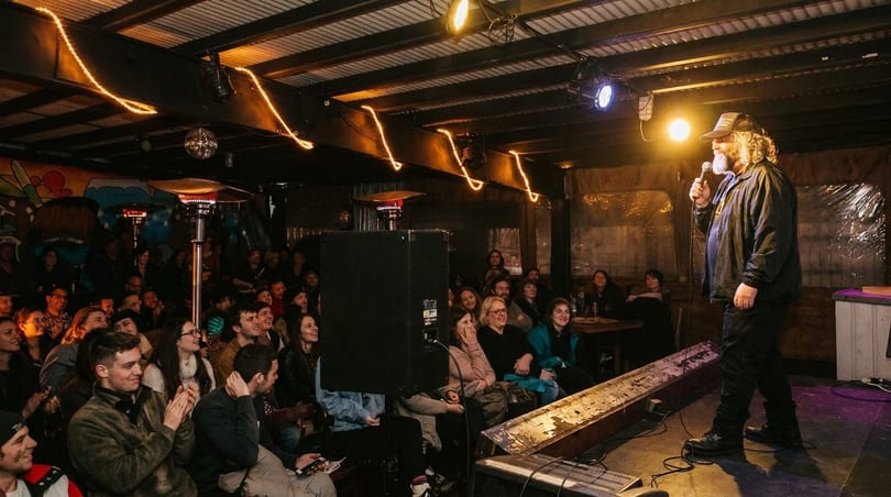 A comedian performs on stage for a laughing audience in a dimly lit venue with string lights.