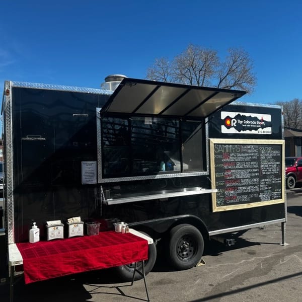 Black food truck "The Colorado Roast" with a menu board and red-covered serving table outdoors.
