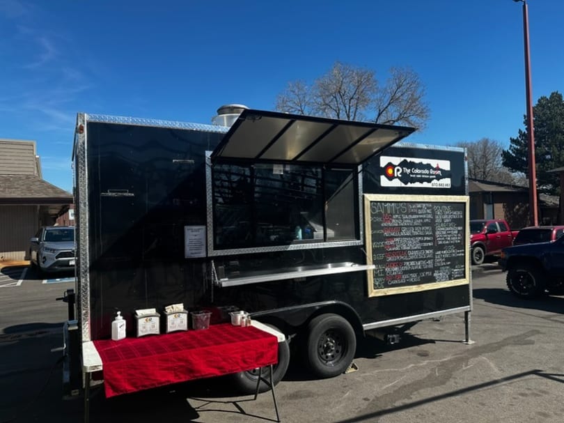 Black food truck "The Colorado Roast" with a menu board and red-covered serving table outdoors.