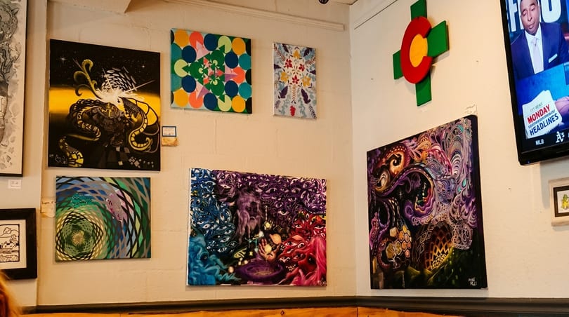A wall covered with various colorful, abstract, and psychedelic paintings, with a TV screen in the corner.