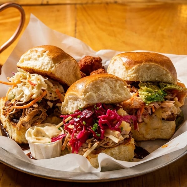Three gourmet sliders with various toppings and a copper mug on a rustic wooden table.