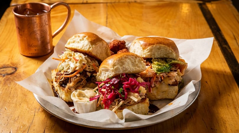 Three gourmet sliders with various toppings and a copper mug on a rustic wooden table.
