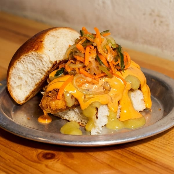 A fried chicken sandwich with orange sauce, green relish, and pickled vegetables on a metal plate.