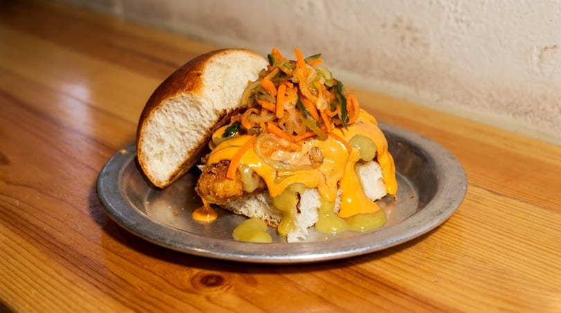 A fried chicken sandwich with orange sauce, green relish, and pickled vegetables on a metal plate.