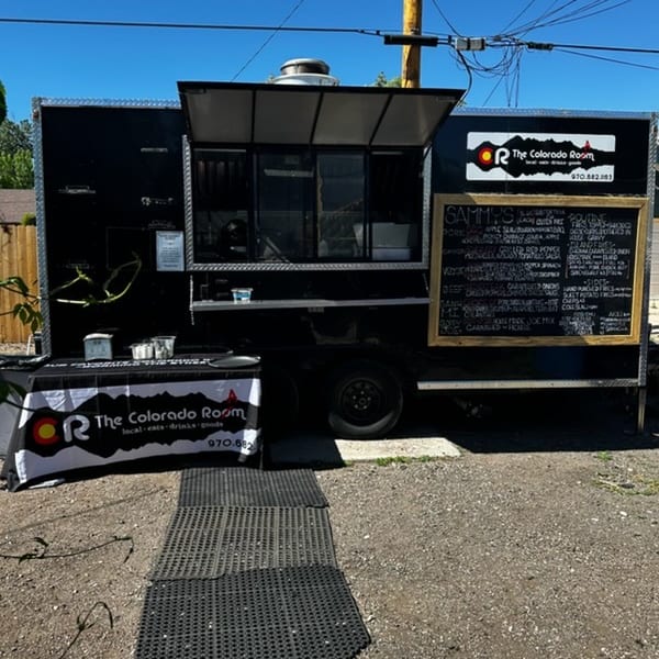 Black food truck for The Colorado Room with a menu board and branded table on a sunny day.