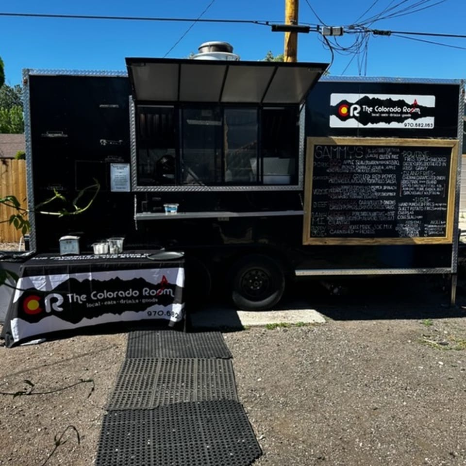 Black food truck for The Colorado Room with a menu board and branded table on a sunny day.