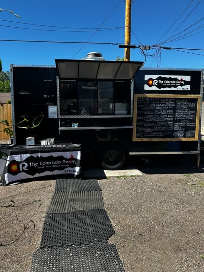 Black food truck for The Colorado Room with a menu board and branded table on a sunny day.