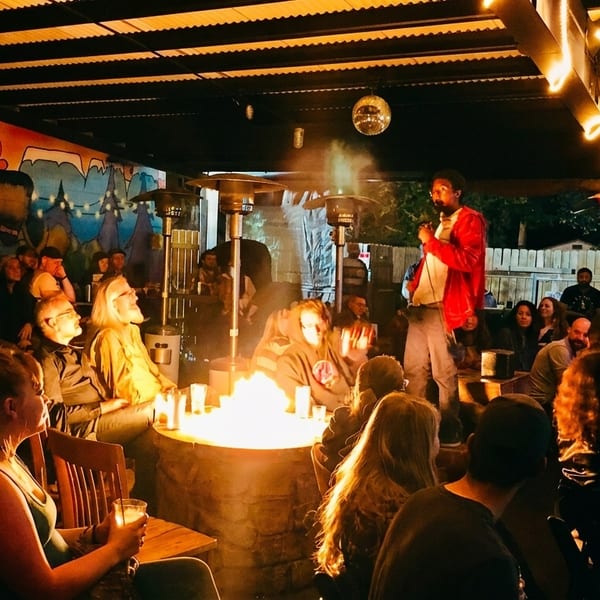 A comedian performs on an outdoor patio with a fire pit, string lights, and a colorful mural.