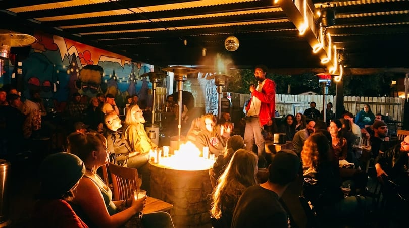 A comedian performs on an outdoor patio with a fire pit, string lights, and a colorful mural.